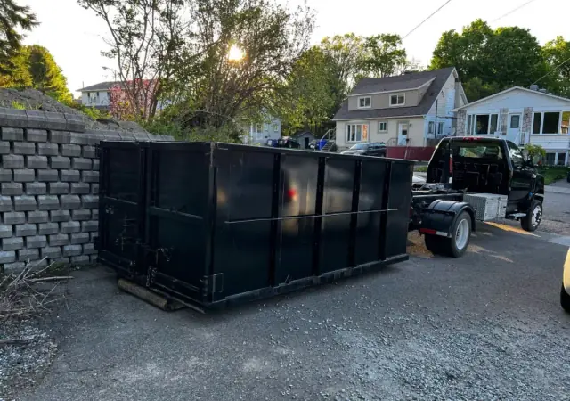 Conteneur noir déposé dans une entrée à côté d’un muret de blocs, le camion à bras qui repart