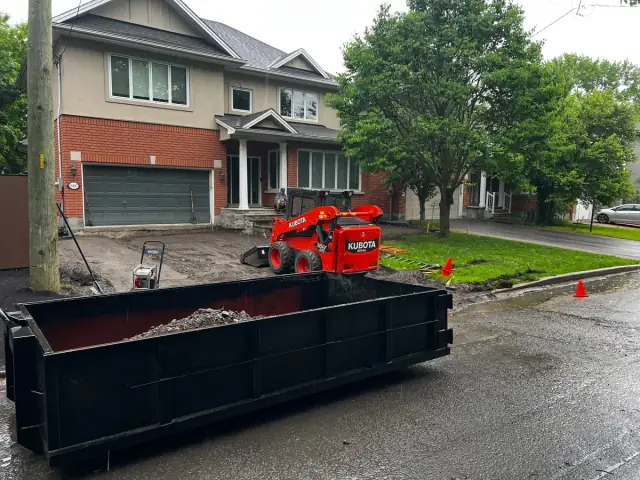 Kubota SSV65 skid steer and a roll-off bin in front of a brick house during a driveway job