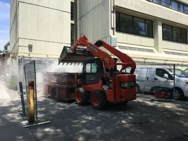 Mini-chargeuse chargeant des débris dans un conteneur, la poussière abattue par un jet d’eau